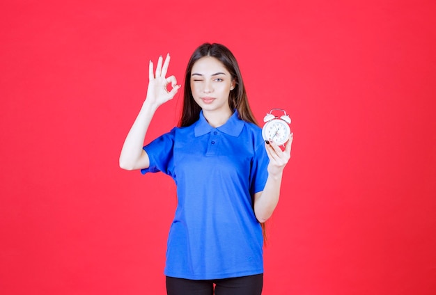 Jeune femme en chemise bleue tenant un réveil et montrant un signe positif de la main