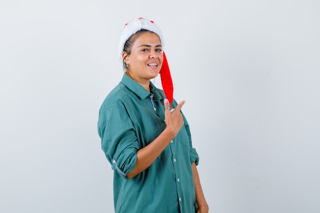 Jeune femme en chemise, bonnet de Noel pointant vers le coin supérieur droit et regardant joyeux, vue de face.