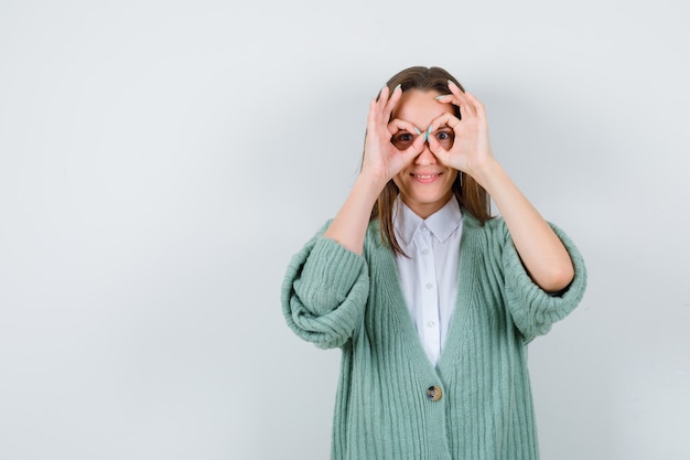 Jeune femme en chemise, cardigan montrant un geste de lunettes et l'air amusé, vue de face.