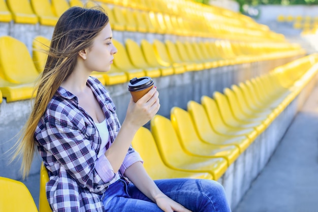 Jeune femme en chemise à carreaux avec gobelet en papier assis au stade