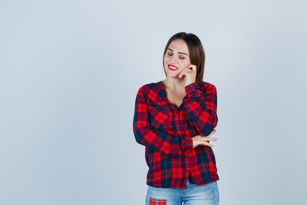 Jeune femme en chemise à carreaux, jeans appuyé sur la joue, souriante, gardant les yeux fermés et l'air heureux, vue de face.