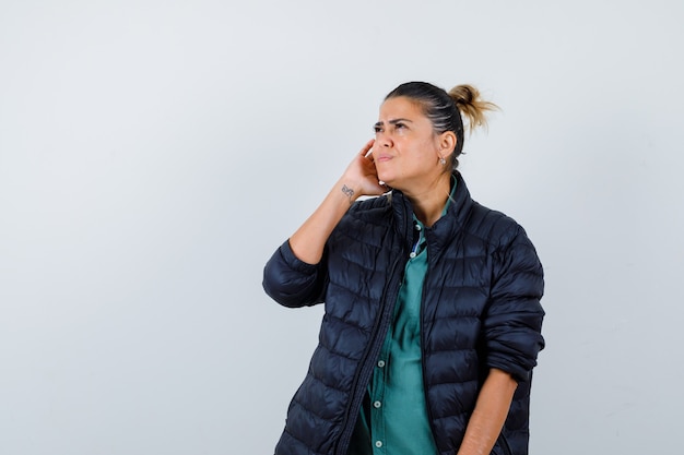 Jeune femme en chemise, doudoune avec la main près de l'oreille, levant les yeux et l'air confus, vue de face.