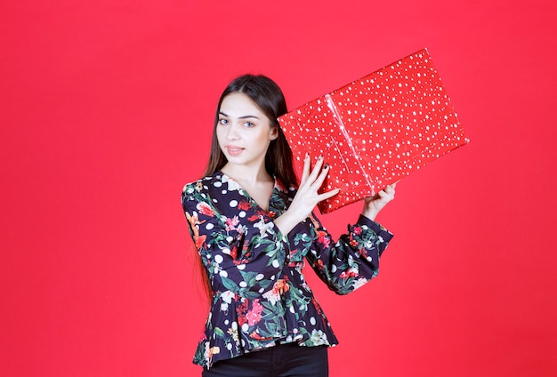 Jeune femme en chemise à fleurs tenant une boîte-cadeau rouge avec des points blancs dessus