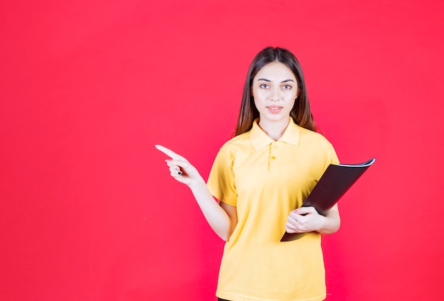 Jeune femme en chemise jaune tenant un dossier noir, pointant et appelant son collègue