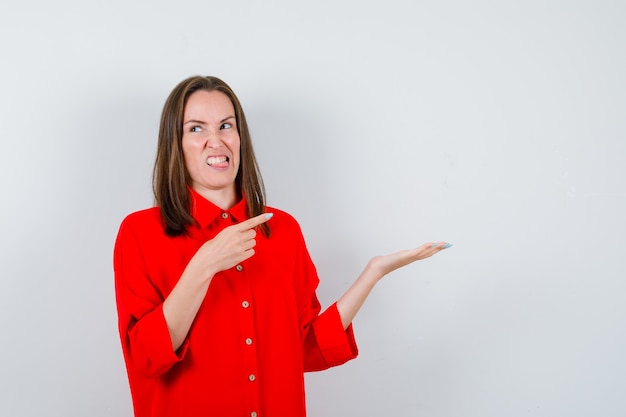 Jeune Femme En Chemisier Rouge Pointant Sur Sa Paume écartée Et L'air Dégoûtée, Vue De Face.