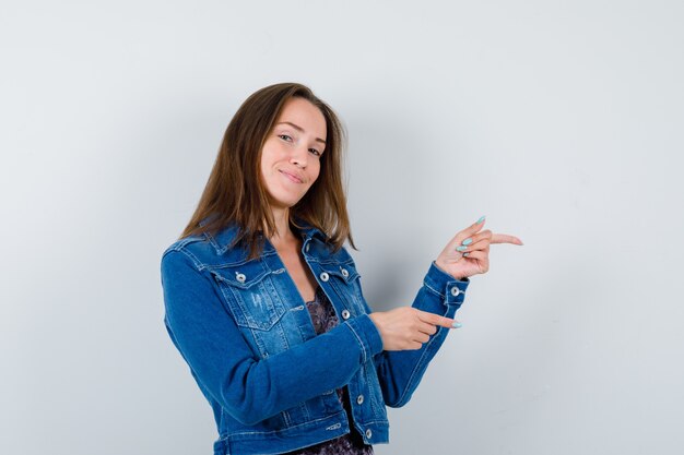 Jeune femme en chemisier, veste en jean pointant vers le côté droit et l'air heureux, vue de face.