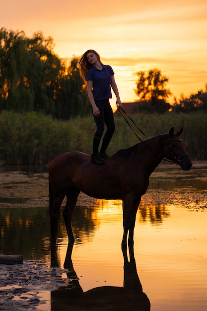 Une jeune femme à cheval sur un lac peu profond. Un cheval court sur l'eau au coucher du soleil. Soin et marche avec le cheval. Force et beauté