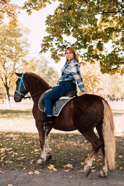 Jeune femme chevauchant un cheval brun posant des promenades en demi-tour dans le parc en automne par une journée ensoleillée