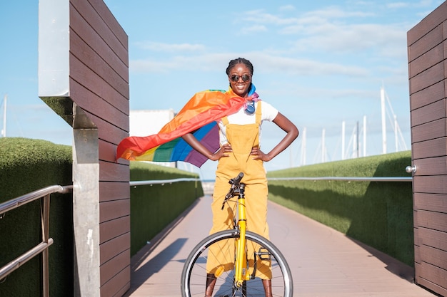 Jeune femme chevauchant un vélo jaune souriant avec le drapeau arc-en-ciel comme cape de super-héros Concept combat de fierté lgtbi