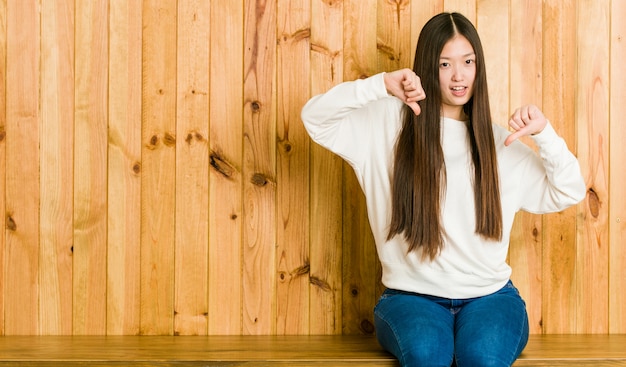 Jeune femme chinoise assise sur une place en bois montrant le pouce vers le bas et exprimant son aversion.