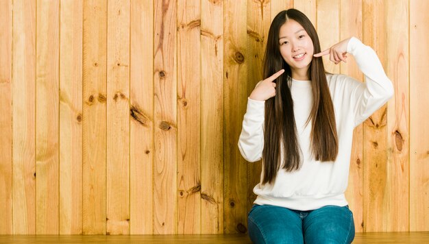 Jeune femme chinoise assise sur une place en bois sourit, pointe ses doigts vers la bouche.