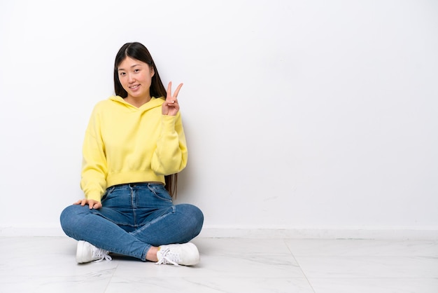 Jeune femme chinoise assise sur le sol isolé sur mur blanc souriant et montrant le signe de la victoire