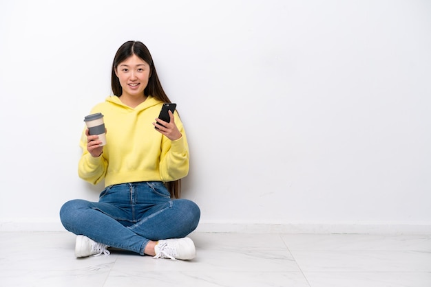 Jeune femme chinoise assise sur le sol isolé sur un mur blanc tenant du café à emporter et un mobile