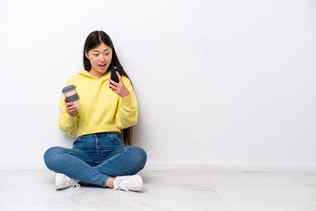 Jeune femme chinoise assise sur le sol isolé sur un mur blanc tenant du café à emporter et un mobile