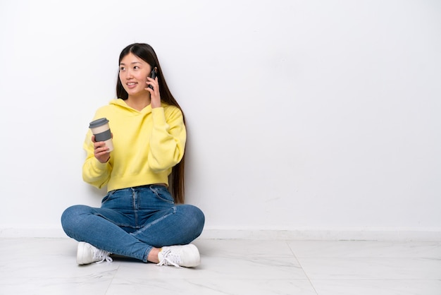 Jeune femme chinoise assise sur le sol isolé sur un mur blanc tenant du café à emporter et un mobile