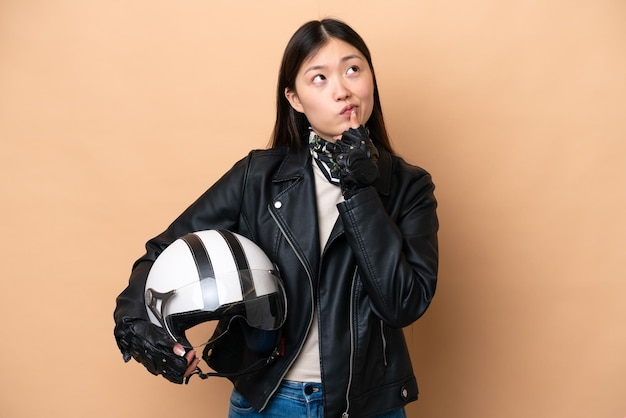 Jeune femme chinoise avec un casque de moto isolé sur fond beige ayant des doutes en levant les yeux