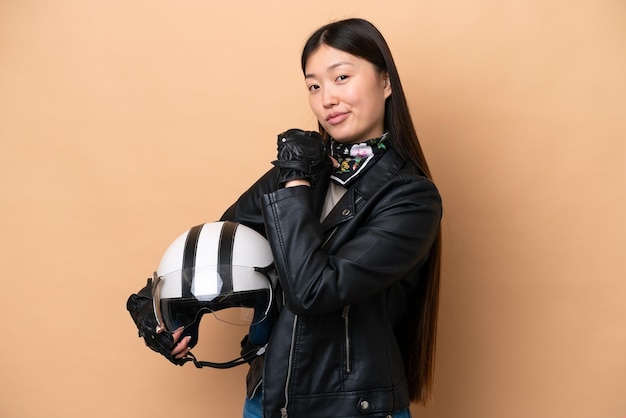 Jeune femme chinoise avec un casque de moto isolé sur fond beige fier et satisfait