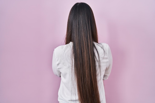 Jeune femme chinoise debout sur fond rose debout en arrière regardant loin avec les bras croisés