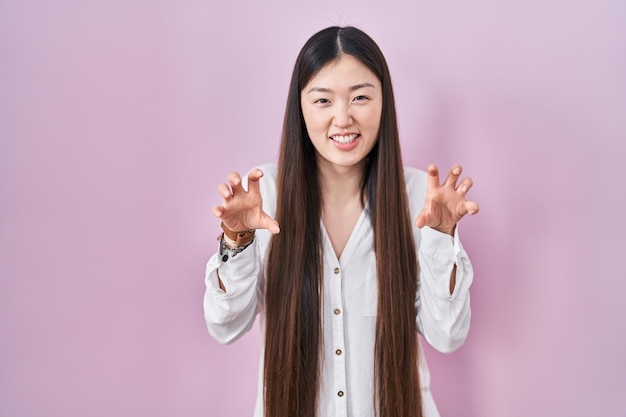 Jeune femme chinoise debout sur fond rose souriant drôle faisant un geste de griffe comme chat expression agressive et sexy