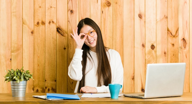 Jeune femme chinoise étudiant sur son bureau excité en gardant le geste ok sur les yeux.