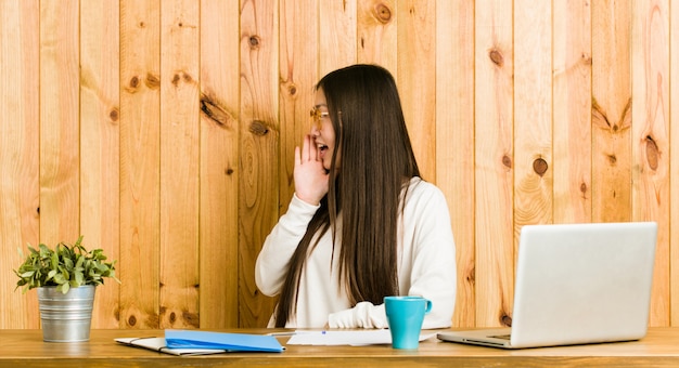 Jeune femme chinoise étudie sur son bureau en criant et en tenant la paume près de la bouche ouverte.