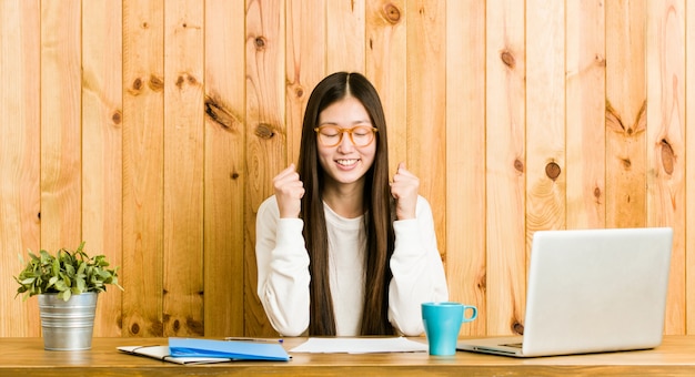 Jeune femme chinoise étudie sur son bureau en levant le poing, se sentant heureuse et réussie.