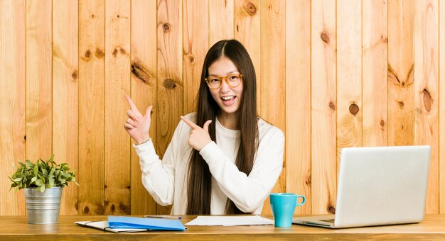 Jeune femme chinoise étudie sur son bureau en montrant quelque chose avec des index, exprimant l'excitation et le désir.