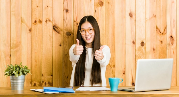 Jeune femme chinoise étudie sur son bureau avec les pouces vers le haut, applaudit à quelque chose, le soutien et le concept de respect.