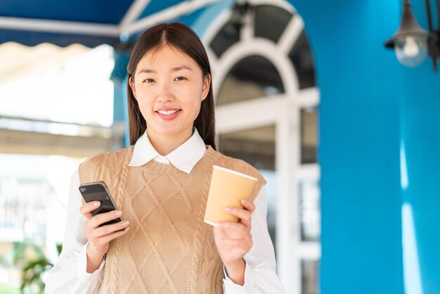 Jeune femme chinoise à l'extérieur à l'aide d'un téléphone portable et tenant un café avec une expression heureuse