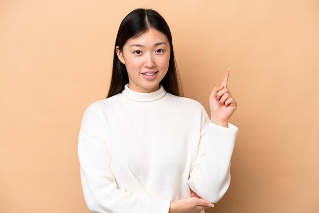 Jeune femme chinoise isolée sur fond beige heureux et pointant vers le haut