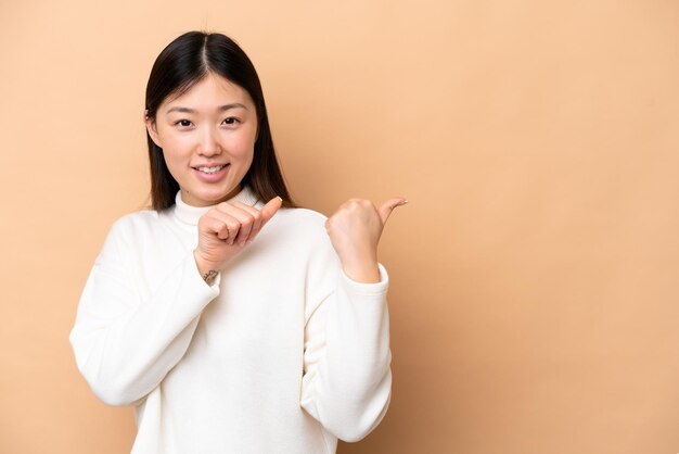 Jeune femme chinoise isolée sur fond beige pointant vers le côté pour présenter un produit