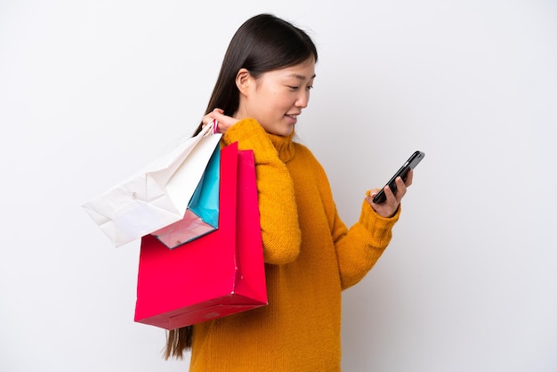 Jeune femme chinoise isolée sur fond blanc tenant des sacs à provisions et écrivant un message avec son téléphone portable à un ami