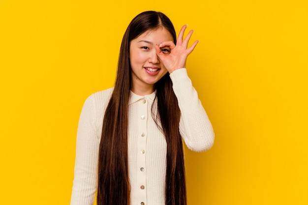 Jeune femme chinoise isolée sur un mur jaune excité en gardant le geste ok sur les yeux.