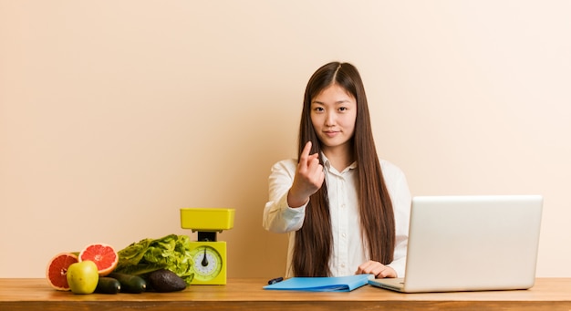 Jeune femme chinoise nutritionniste travaillant avec son ordinateur portable en vous montrant du doigt comme si vous vous y approchiez.