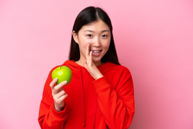 Jeune femme chinoise avec une pomme isolée sur fond rose chuchotant quelque chose