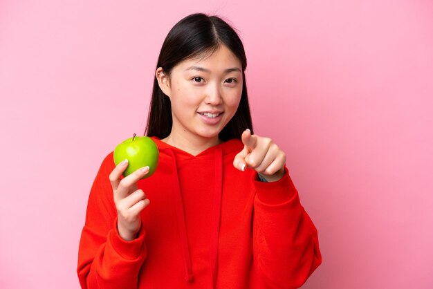 Jeune femme chinoise avec une pomme isolée sur fond rose pointe le doigt vers vous avec une expression confiante