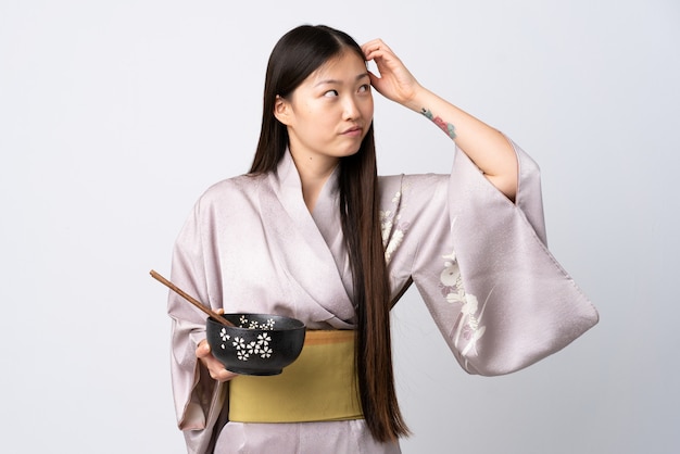 Jeune femme chinoise portant un kimono sur un mur blanc isolé ayant des doutes et avec une expression du visage confuse tout en tenant un bol de nouilles avec des baguettes