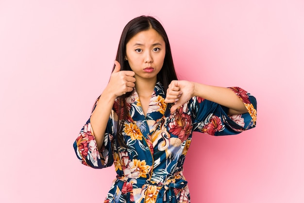 Jeune femme chinoise portant un pyjama kimono isolé montrant les pouces vers le haut et les pouces vers le bas, difficile de choisir le concept