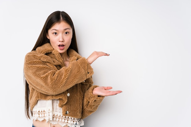 Jeune femme chinoise posant dans un mur blanc isolé choqué et étonné tenant un espace de copie entre les mains.