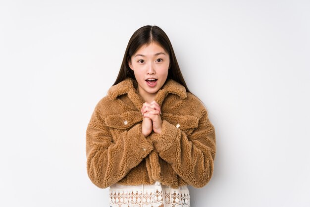 Jeune femme chinoise posant dans un mur blanc isolé priant pour la chance, étonné et ouvrant la bouche en regardant vers l'avant.