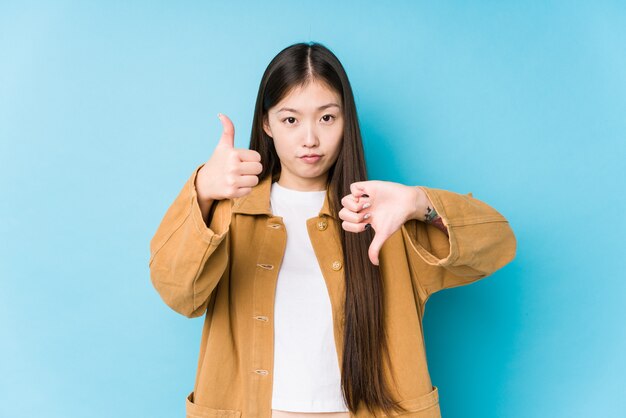 Jeune femme chinoise posant dans un mur bleu isolé montrant les pouces vers le haut et les pouces vers le bas, difficile de choisir le concept