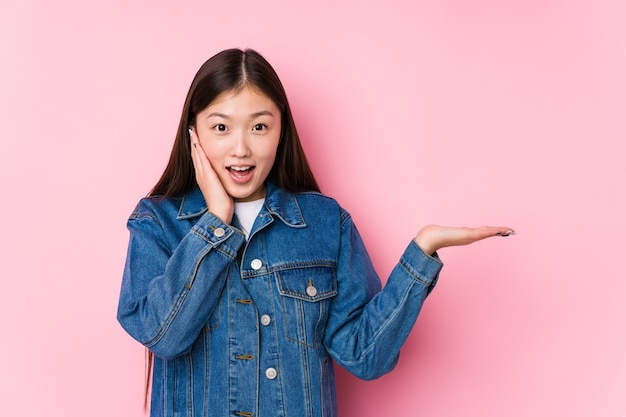 Jeune femme chinoise posant dans un rose isolé détient copie espace sur une paume, garder la main sur la joue