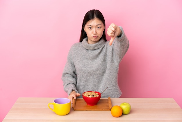 Jeune femme chinoise prenant son petit déjeuner dans une table montrant le pouce vers le bas avec une expression négative