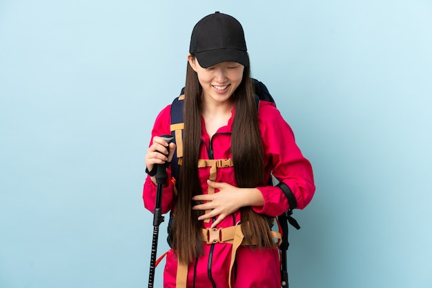 Jeune femme chinoise avec sac à dos et bâtons de randonnée sur un mur bleu isolé souriant beaucoup