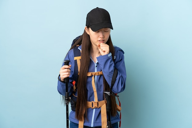 Jeune femme chinoise avec sac à dos et bâtons de randonnée sur un mur bleu isolé toussant beaucoup