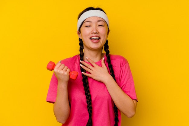 Jeune femme chinoise de sport isolée sur jaune éclate de rire en gardant la main sur la poitrine.