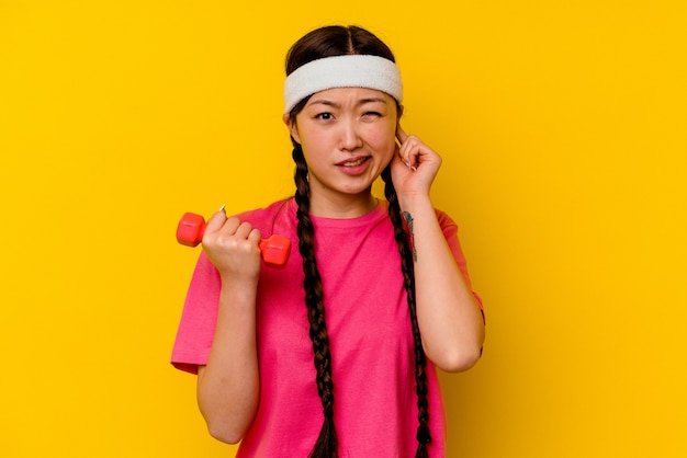 Jeune femme chinoise de sport isolée sur un mur jaune couvrant les oreilles avec les mains
