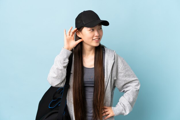 Jeune femme chinoise sport avec sac de sport sur mur bleu isolé écouter quelque chose en mettant la main sur l'oreille