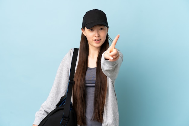 Jeune femme chinoise sport avec sac de sport sur mur bleu isolé montrant et levant un doigt