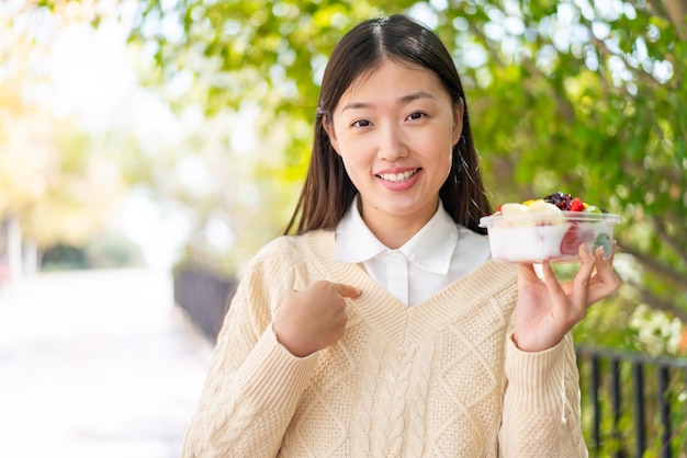 Jeune femme chinoise tenant un bol de fruits à l'extérieur avec une expression faciale surprise